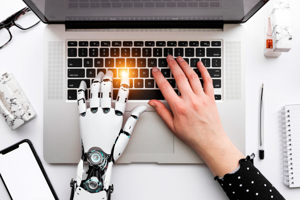 Dwie dłonie ułożone na klawiaturze laptopa; jedna dłoń ludzka, druga robota; w tle na stole przedmioty biurowe, telefon i okulary.