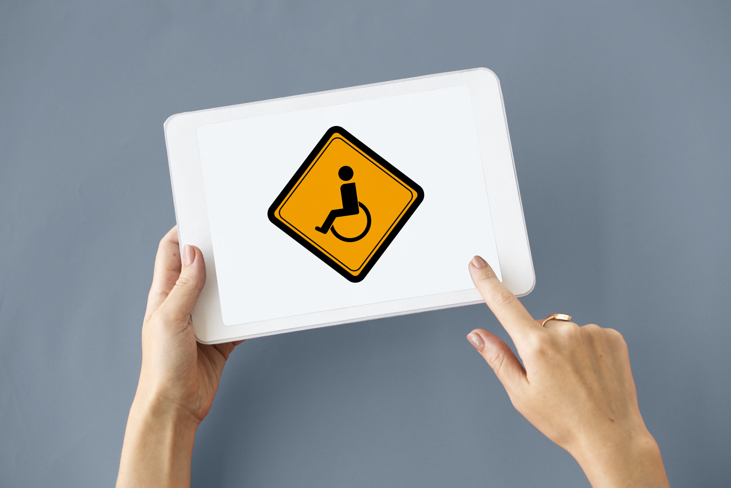tablet z ikoną osoby niepełnosprawnej, obok ręka, która go trzyma a druga wskazuje na niego